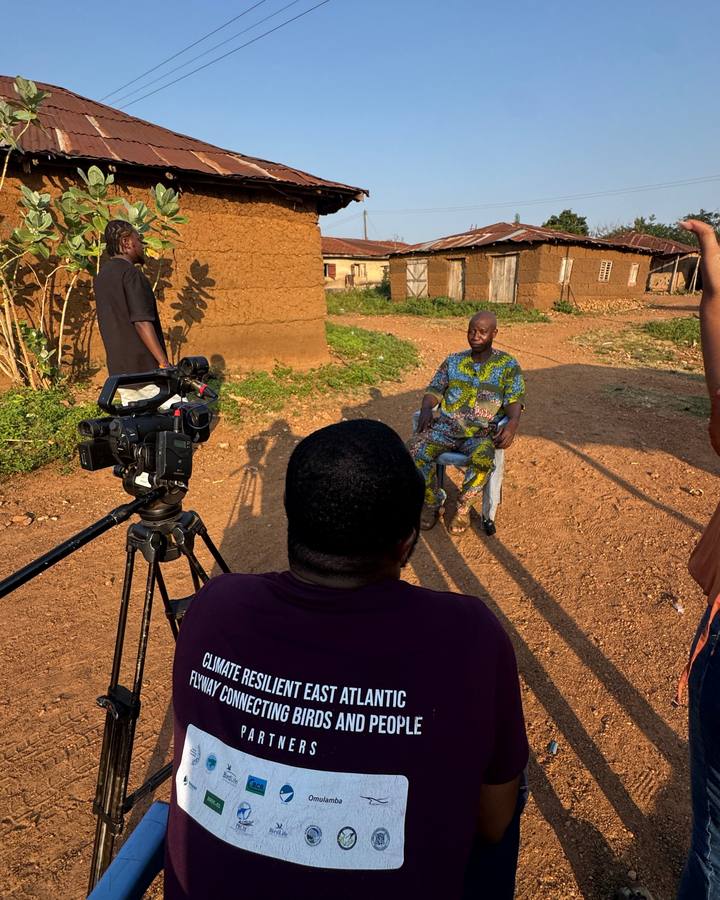 A camera crew filming an interview with a former ranger outside mud-brick houses in Olokemeji